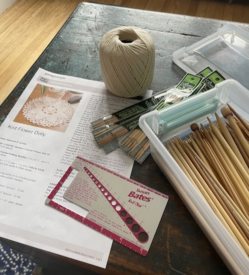 a box of dpns, a doily pattern, crochet thread, and a needle gauge (all on the coffee table)
