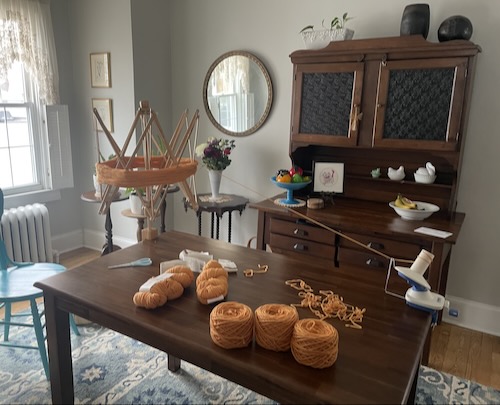 the dining table with a swift attached to one end and the ball winder attached to the other. pumpkin colored yarn is suspended between them and several wound cakes of the same are piled on the table. two skeins remain unwound